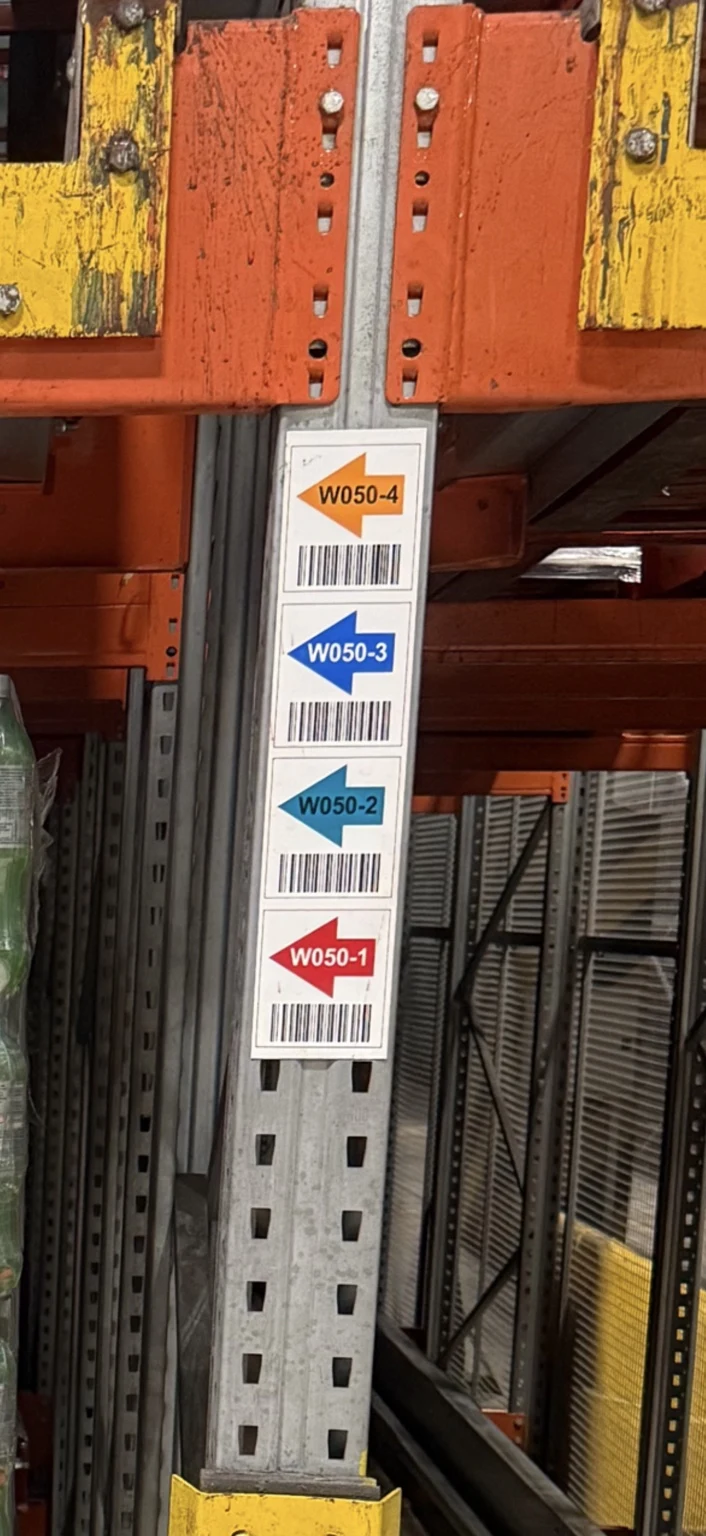 Colour-coded location labels with directional arrows and barcodes on a shuttle rack upright